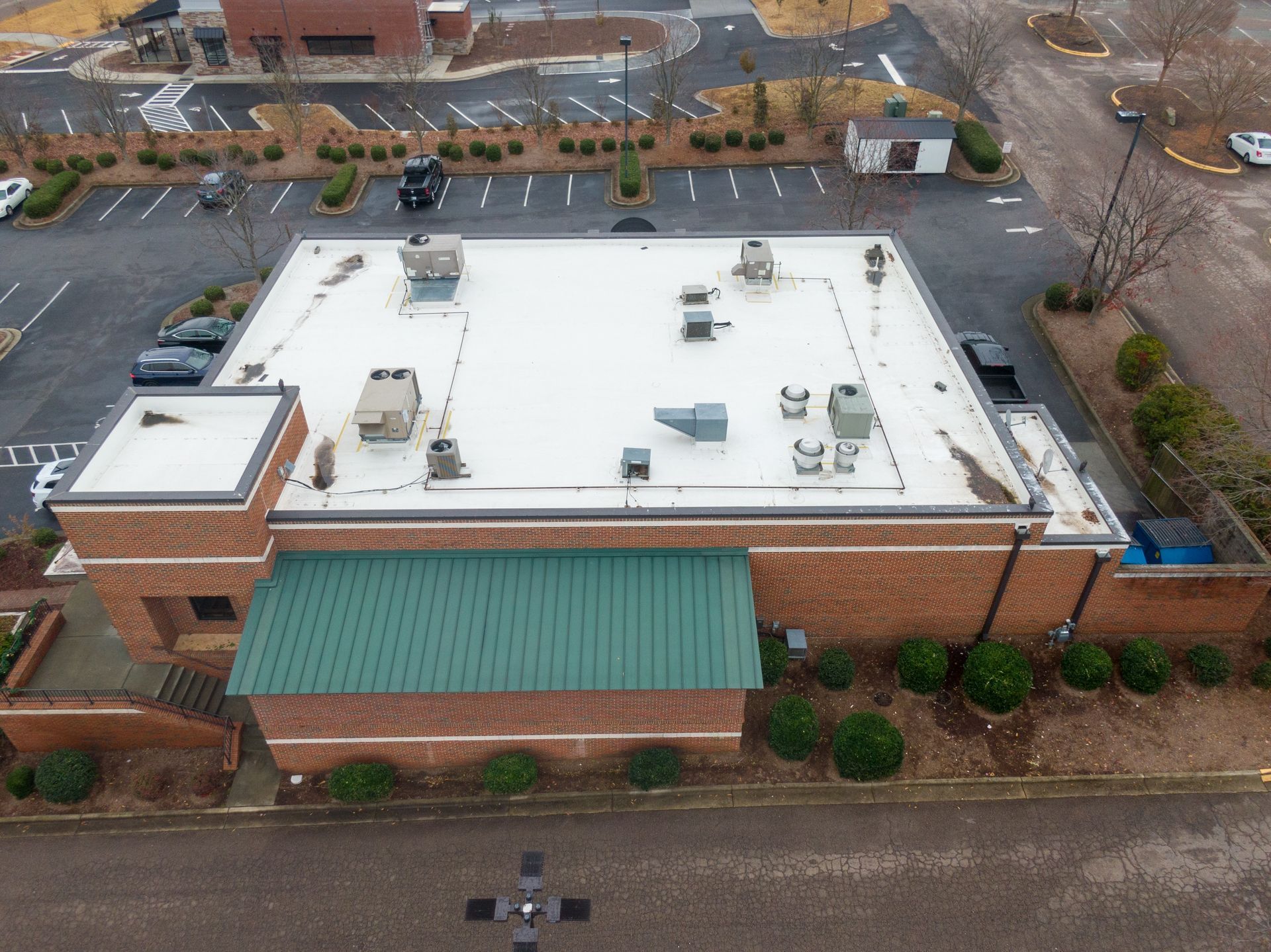 Aerial view of a single-story brick building with a flat white roof. A green awning extends from the front. A parking lot surrounds it.