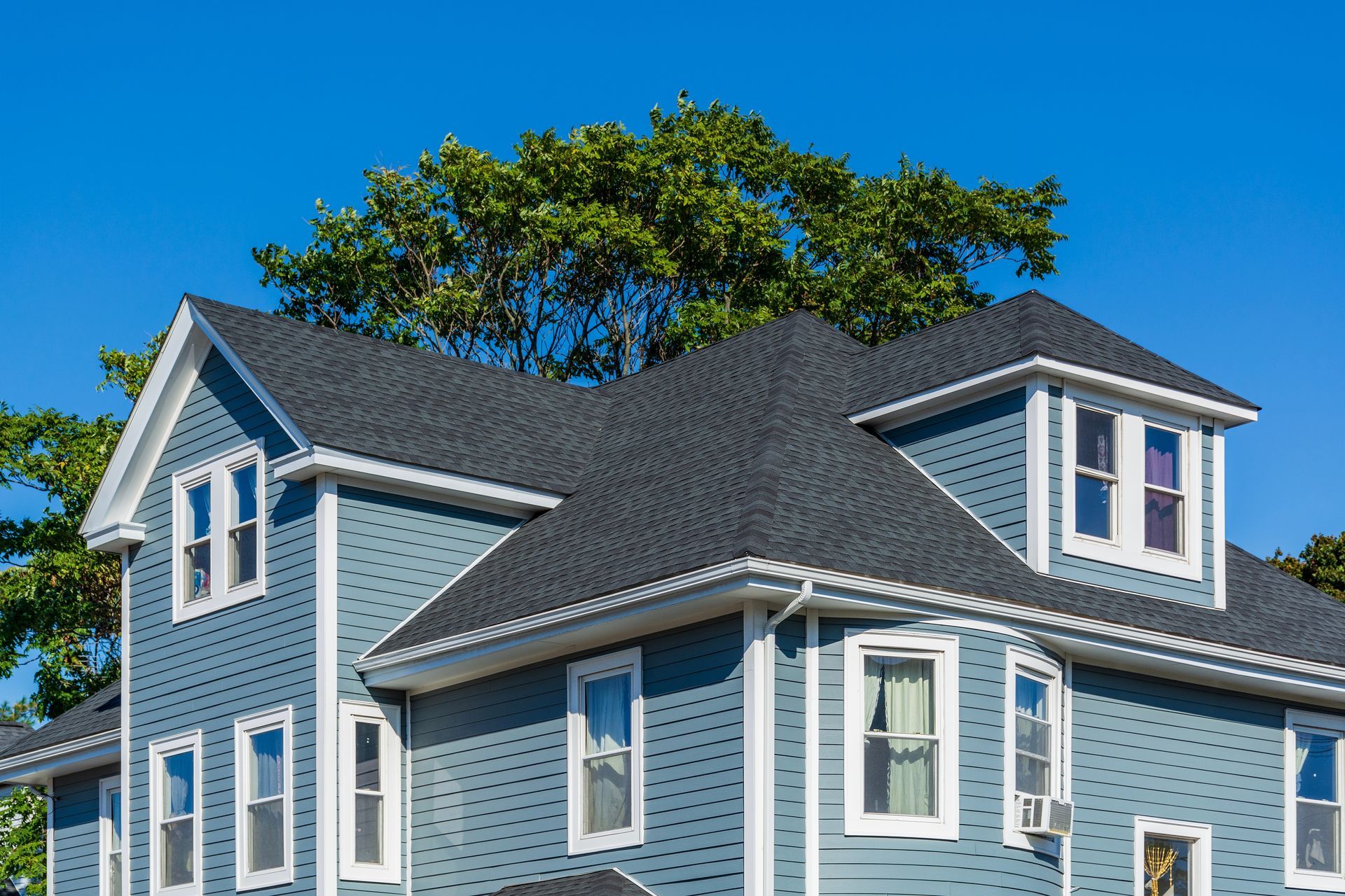 Blue two-story house with dark gray roof against a clear blue sky. Tree branches in the background.
