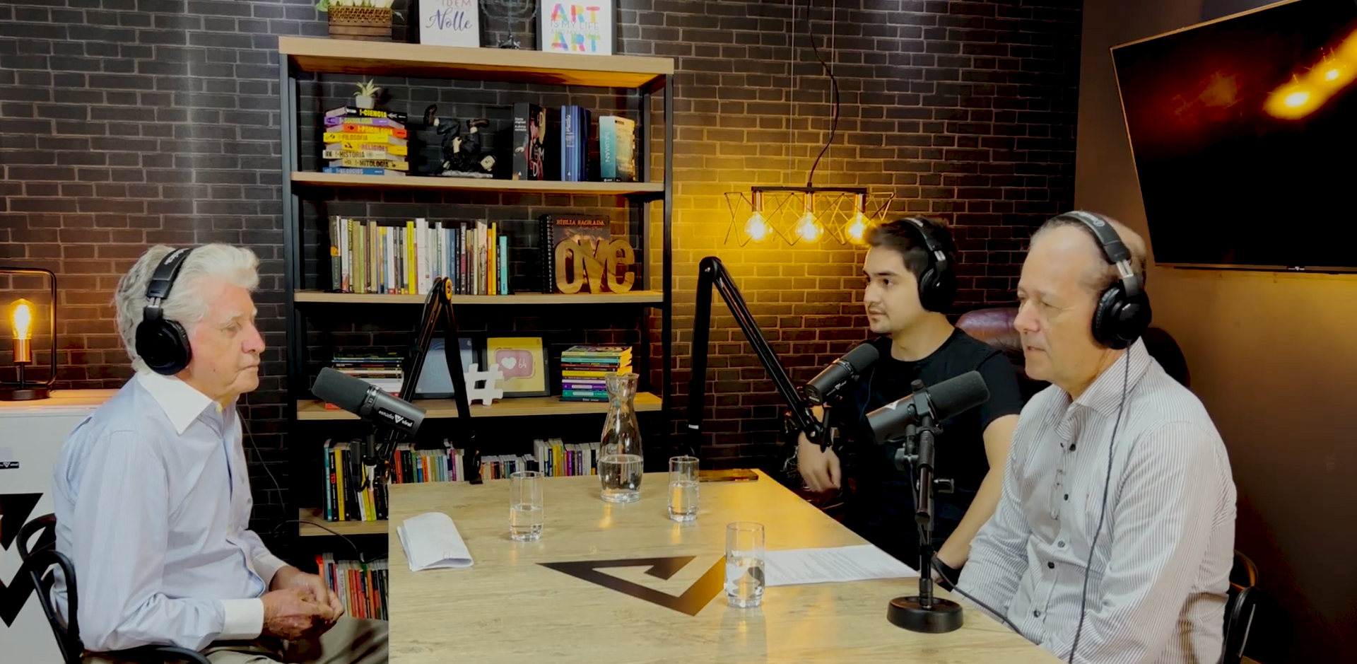Três pessoas usando fones de ouvido sentam-se à mesa com microfones em uma sala com paredes de tijolos durante a gravação de um podcast.