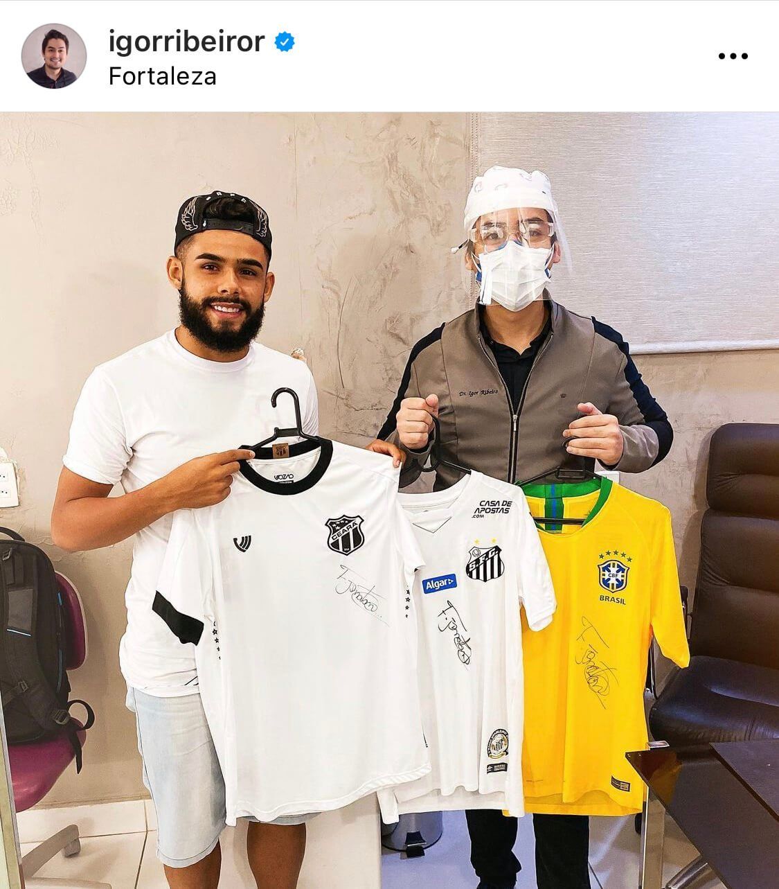 Homem segurando camisas autografadas, uma amarela do Brasil e duas brancas do Santos. Outro homem usa protetor facial.