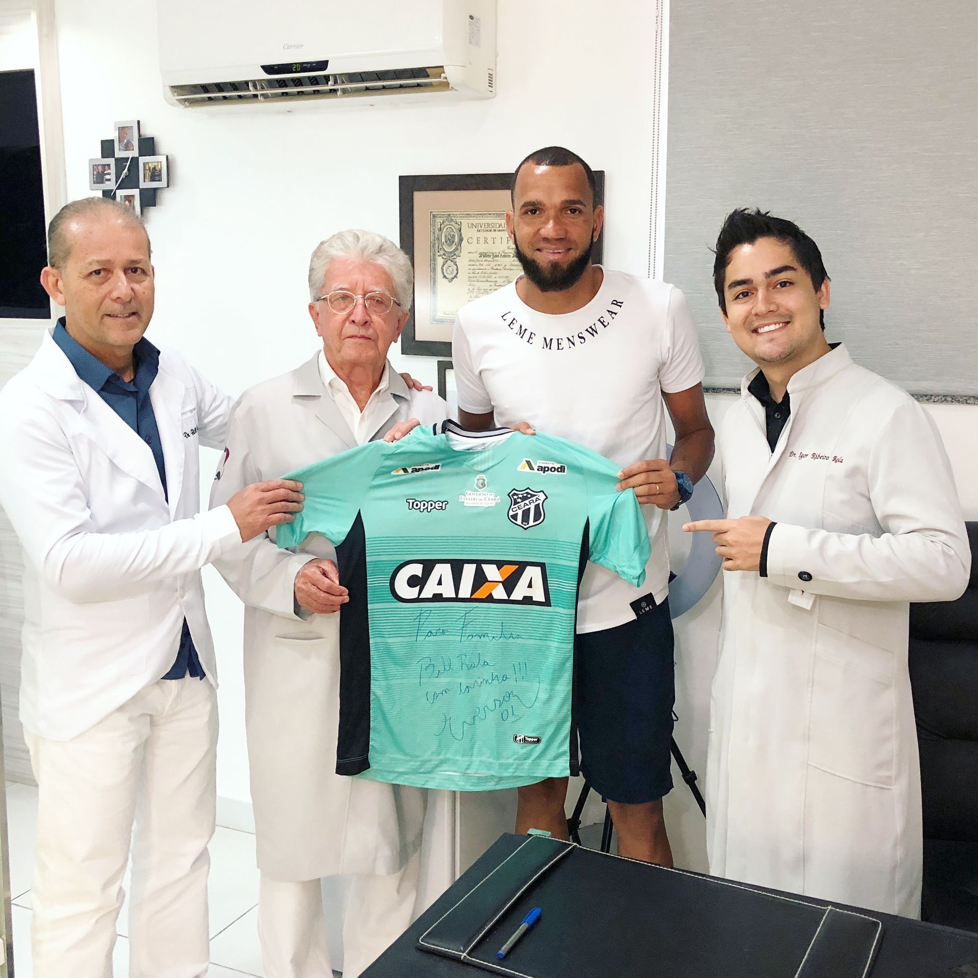 Em um escritório, três profissionais de jaleco branco e um homem de camiseta branca seguram uma camisa de futebol verde-clara autografada.