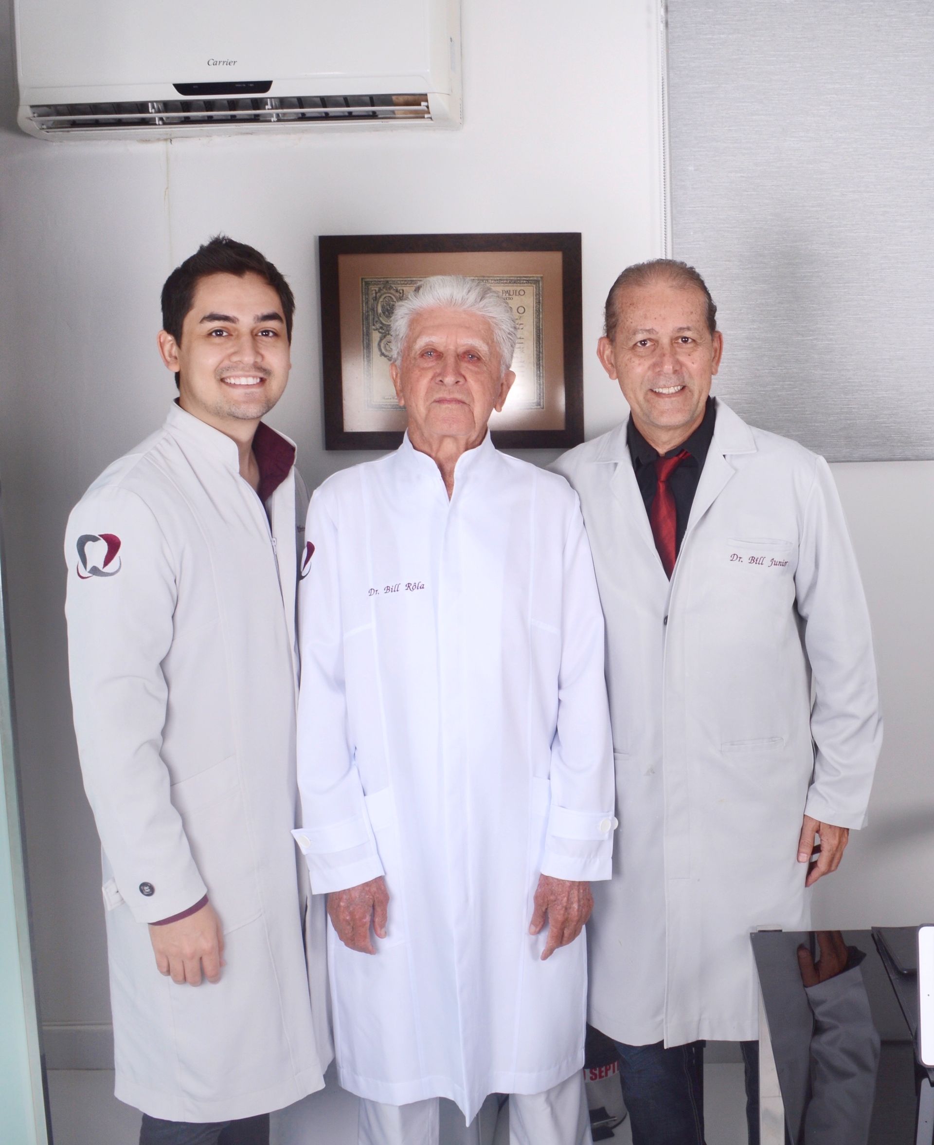 Três pessoas vestindo jalecos brancos profissionais estão juntas, sorrindo, em um escritório bem iluminado.
