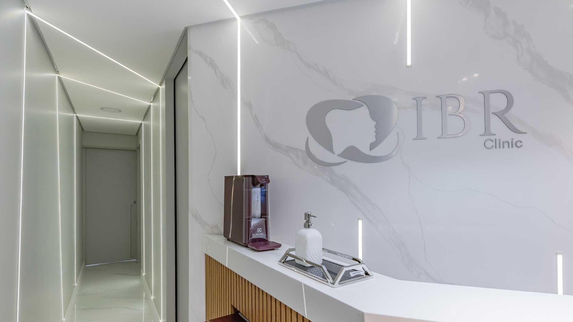 Recepção moderna de clínica odontológica com parede de mármore branco, logotipo, balcão e corredor com iluminação linear.