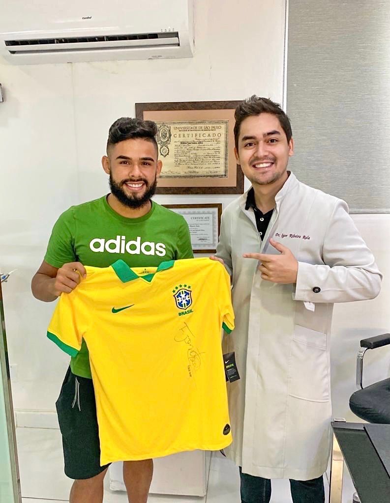 Homem segurando uma camisa da seleção brasileira de futebol ao lado de outro homem, ambos sorrindo em um ambiente interno.