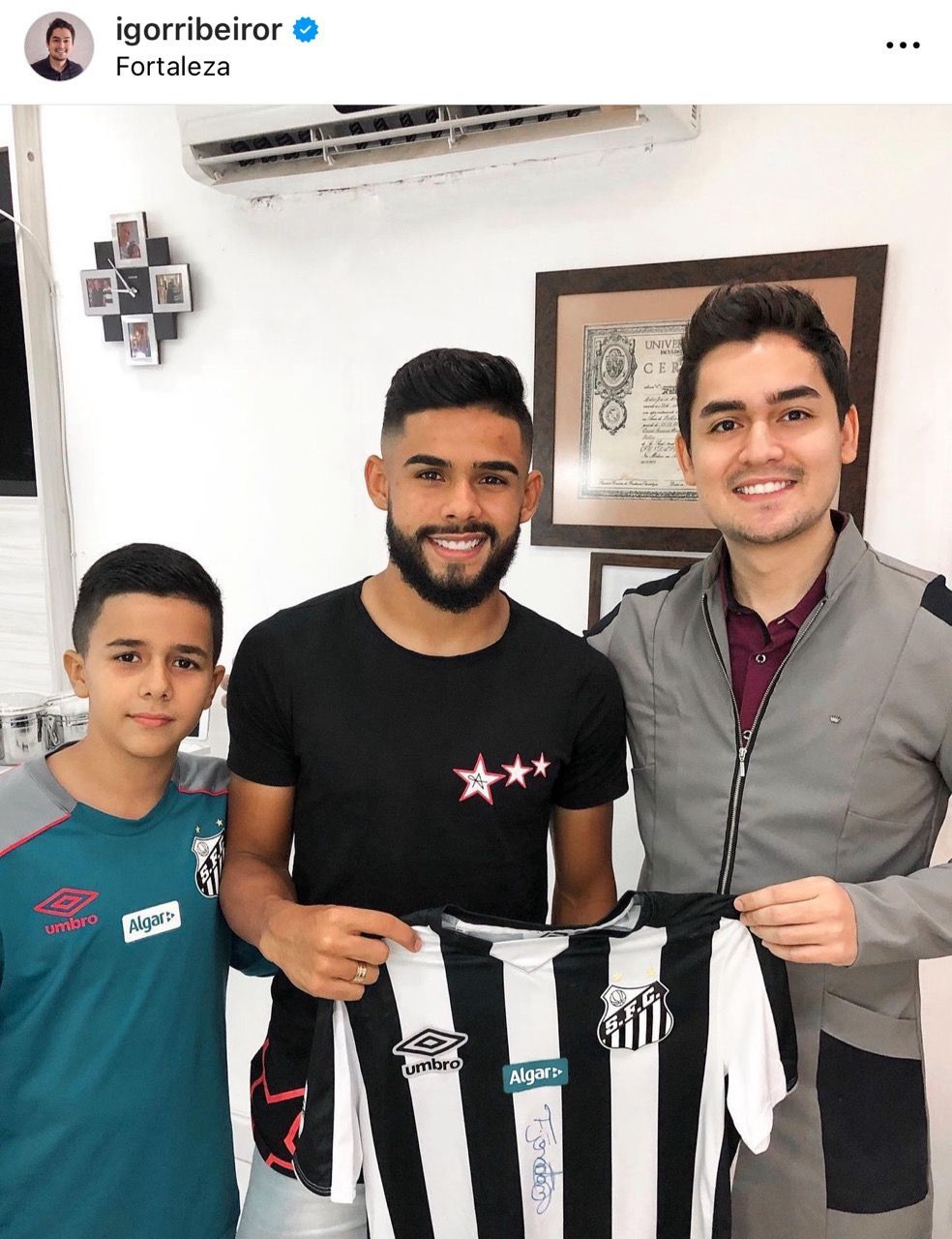 Homem segurando uma camisa de futebol, ladeado por duas pessoas, sorrindo dentro de um ambiente fechado.