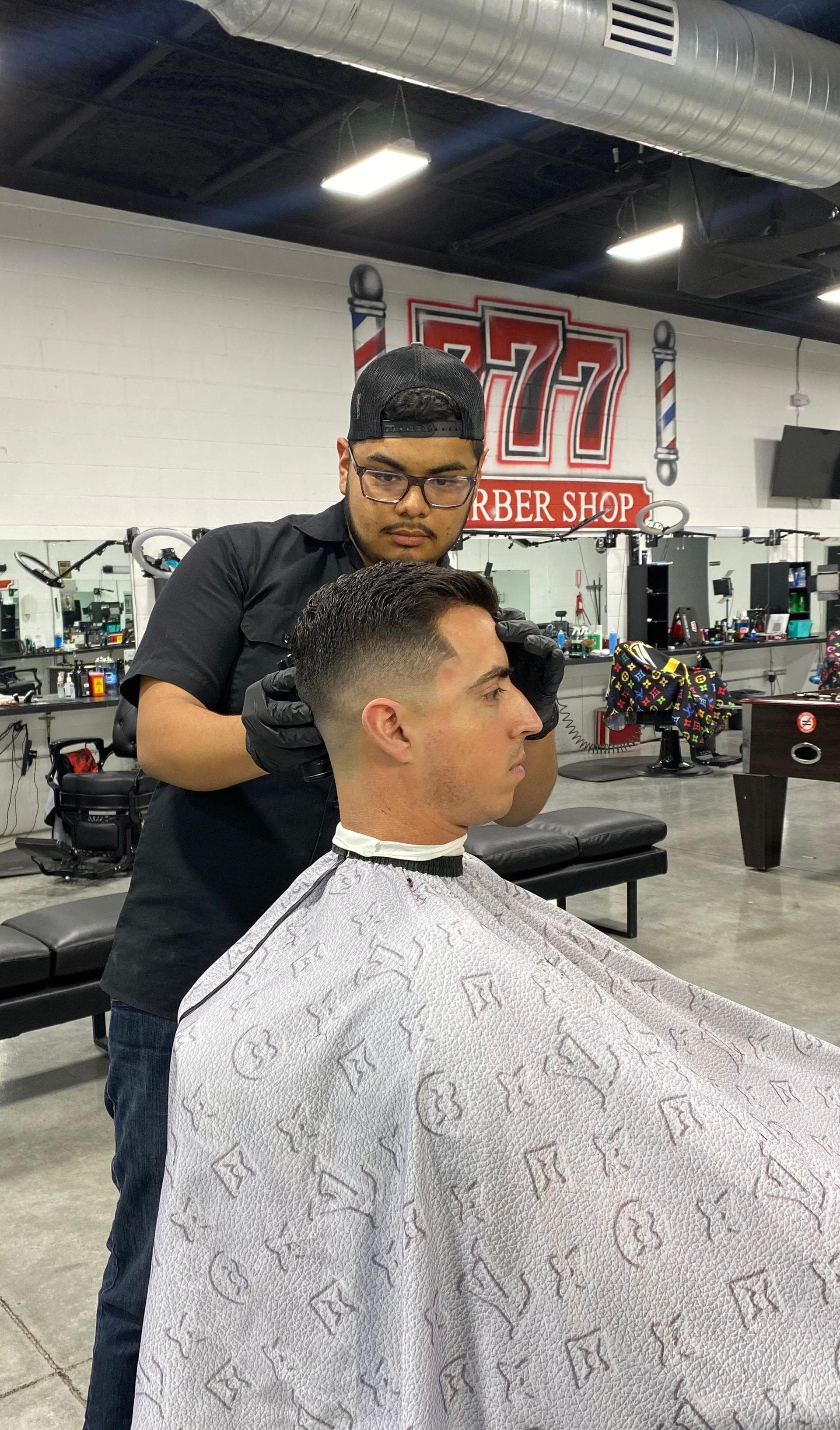 Barber giving a client a haircut in a shop. Client wears a cape, barber in black clothing.
