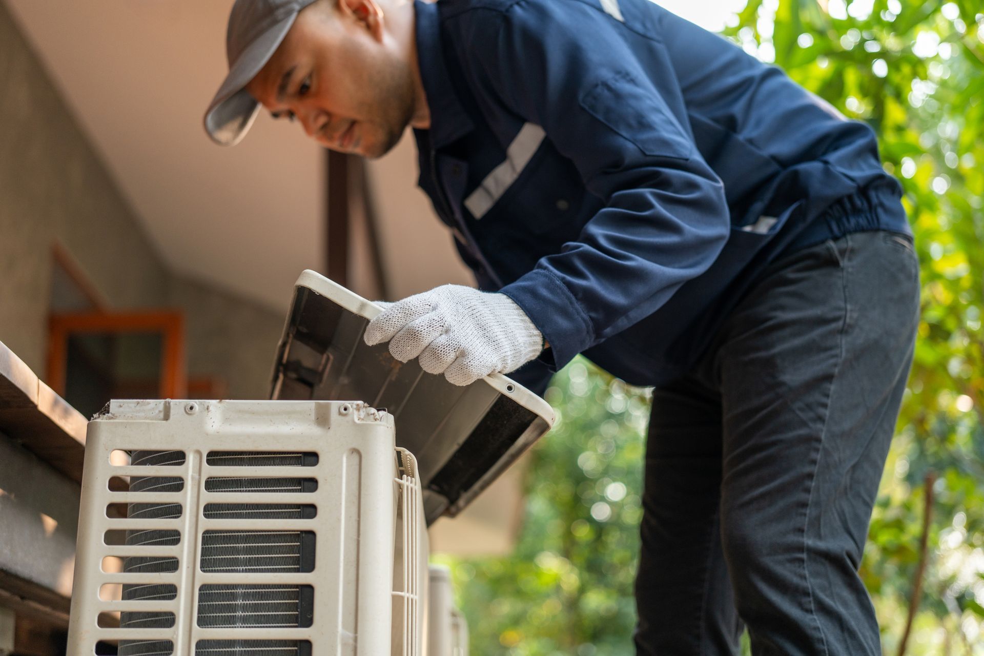 Technician inspecting outdoor HVAC unit beside a building, wearing blue coveralls and gloves.