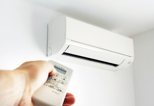 Hand holding a remote aimed at a white wall-mounted air conditioner turning it on