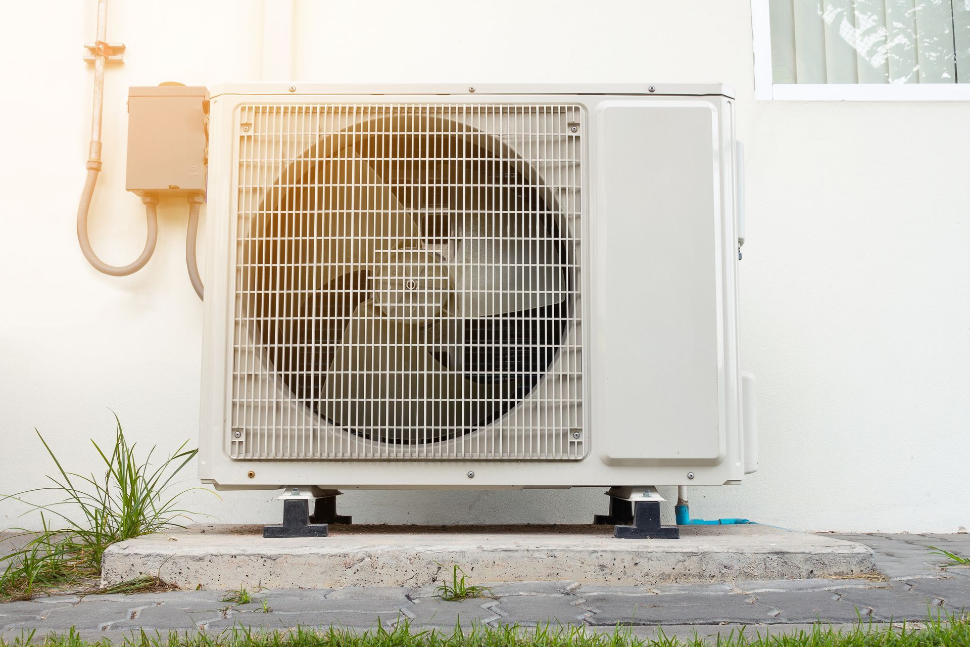 Outdoor air conditioner unit against a white wall, with a plant and wall light nearby