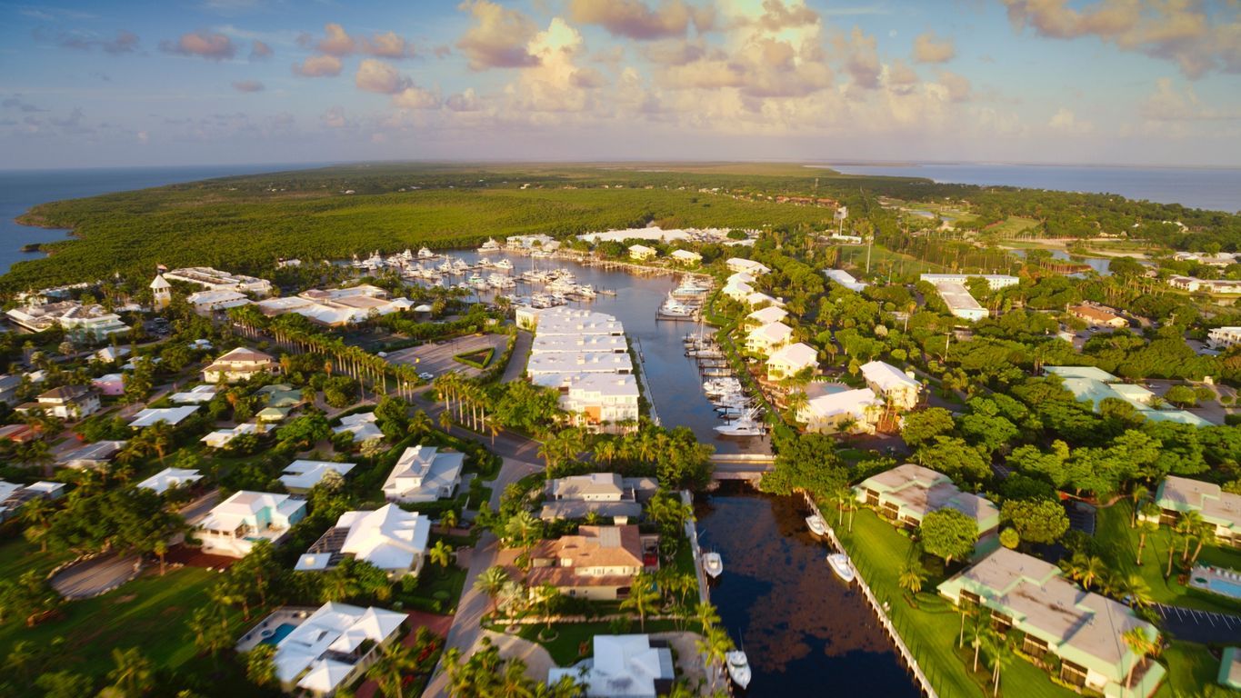 Aerial view of a coastal neighborhood with canals, white-roofed homes, and lush green trees under a sunset sky
