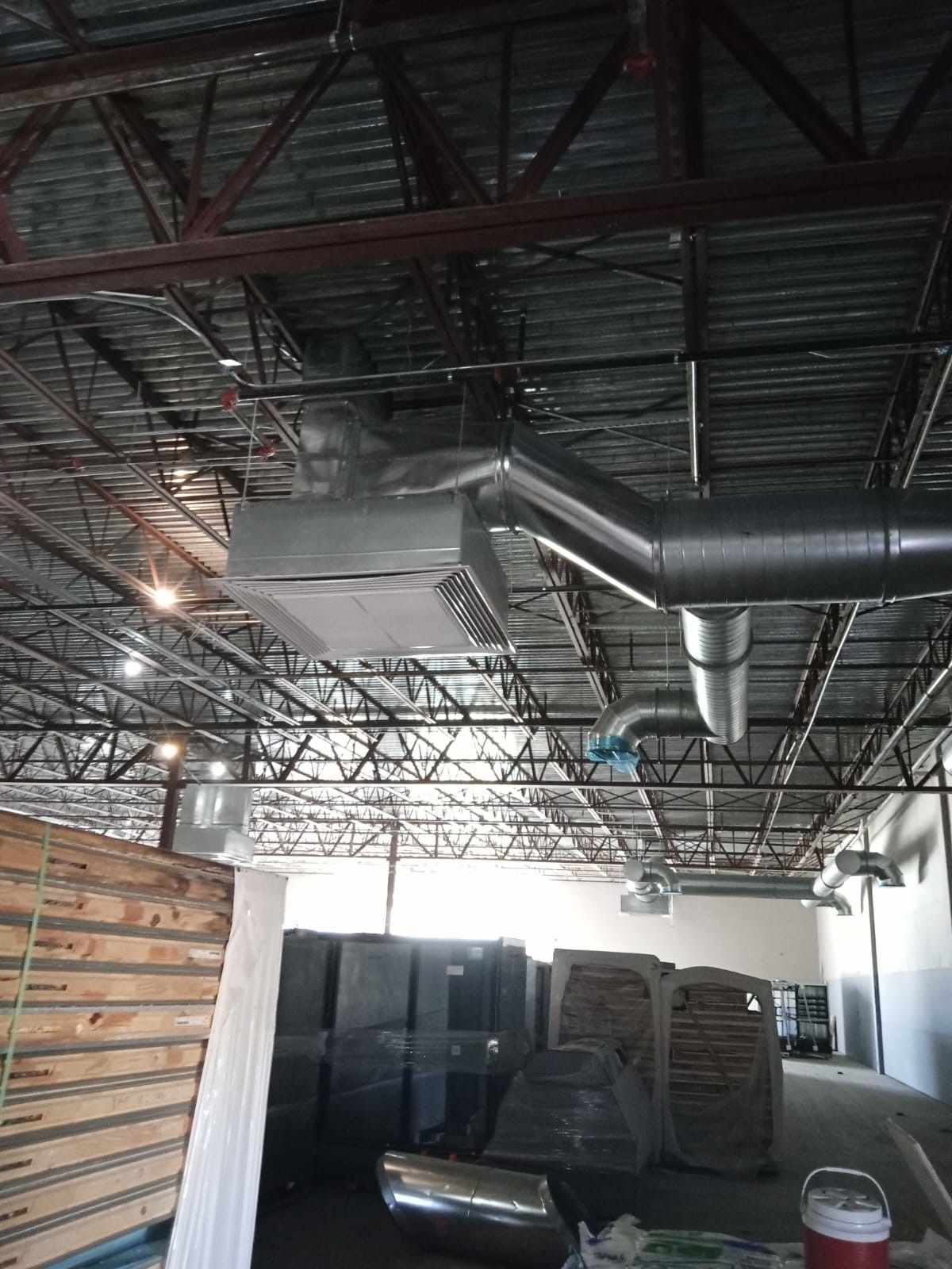 Industrial warehouse interior with exposed steel roof trusses, ductwork, stacked equipment, and wood paneling.
