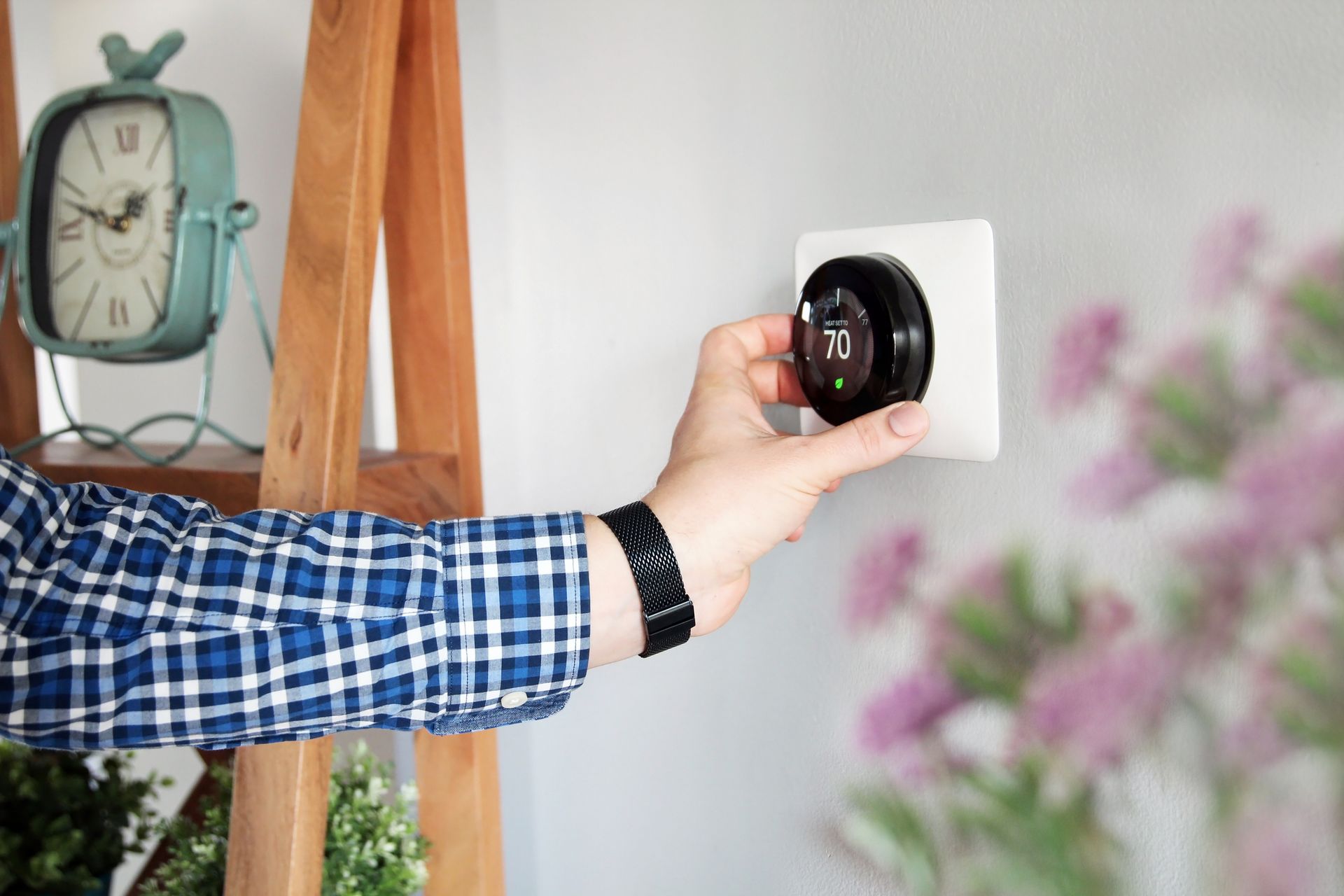 Hand turning a wall thermostat in a bright room, with a clock and wooden shelf nearby