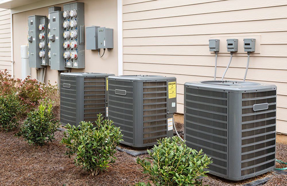 Three outdoor HVAC units beside a beige house with electrical meters and shrubs