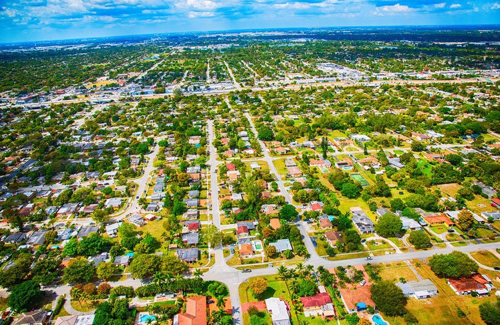 Aerial view of a suburban neighborhood with grid-like streets, colorful houses, and green trees under a blue sky