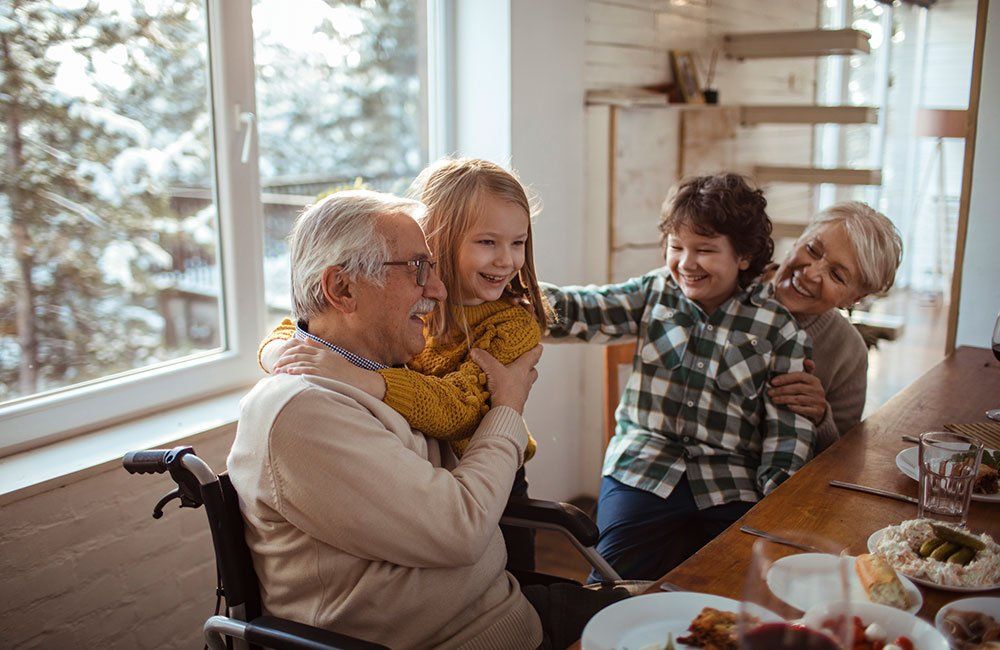 Family gathered at a dining table, smiling and embracing in a bright room by a window.