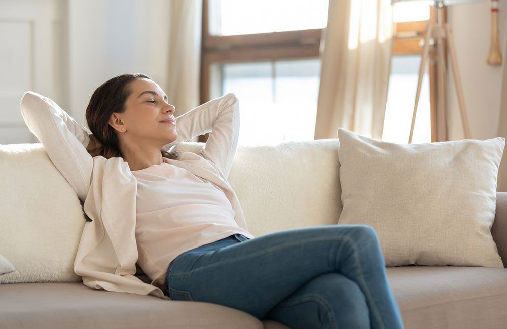 Woman reclining on a beige couch with hands behind her head in a bright living room