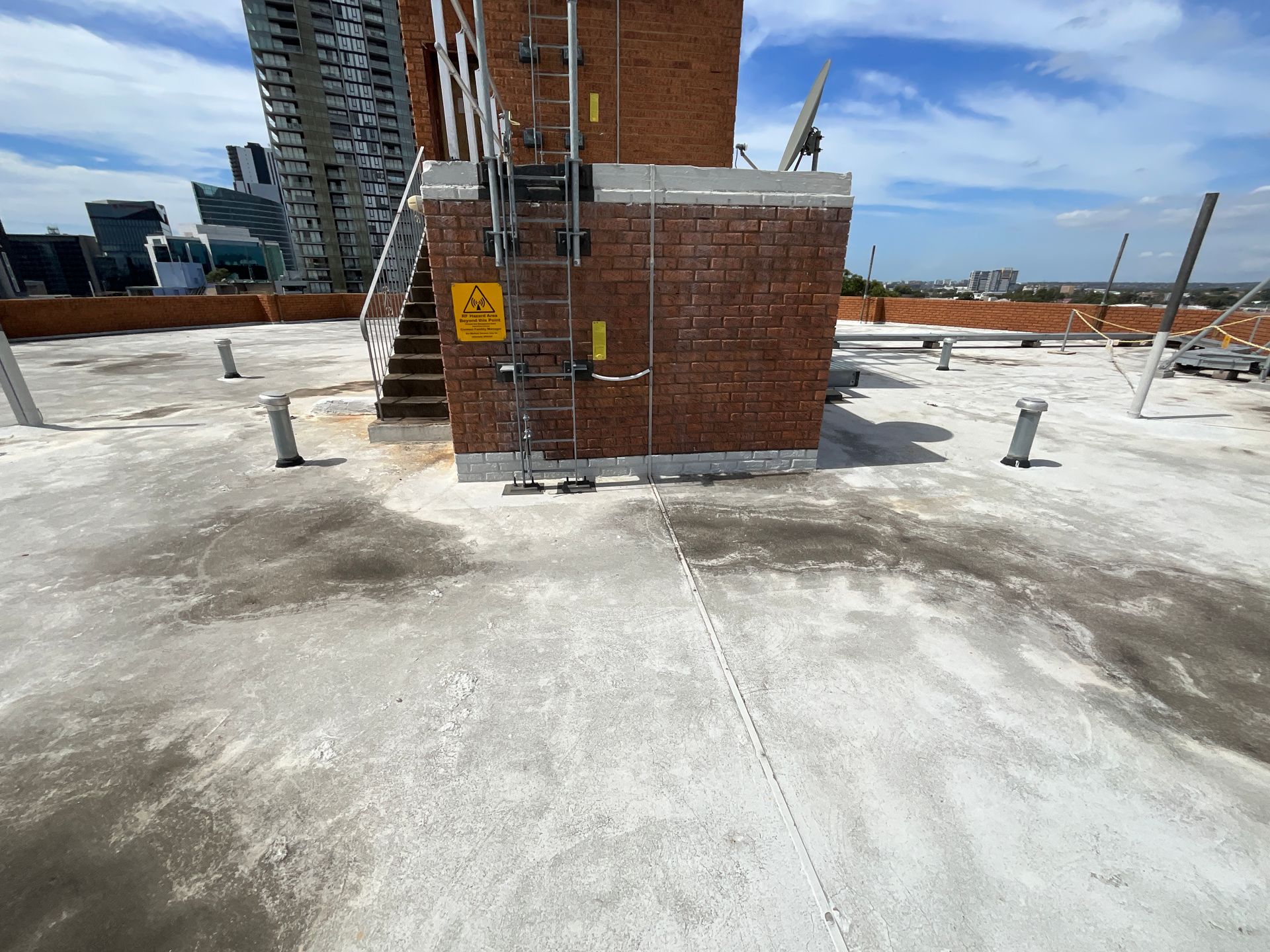 Rooftop with a brick utility structure, ladder, pipes, and city buildings in the background