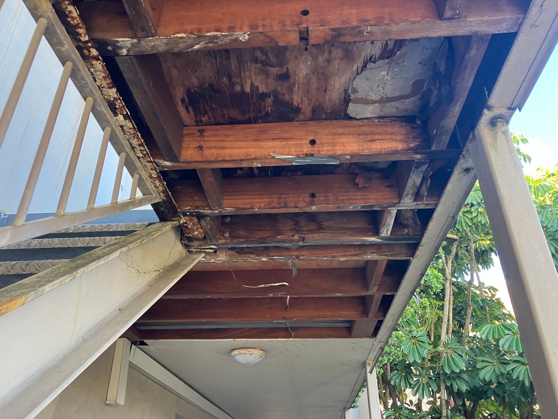 Under-stair view of exposed wooden beams and rusty underside, with a white railing and ceiling light below