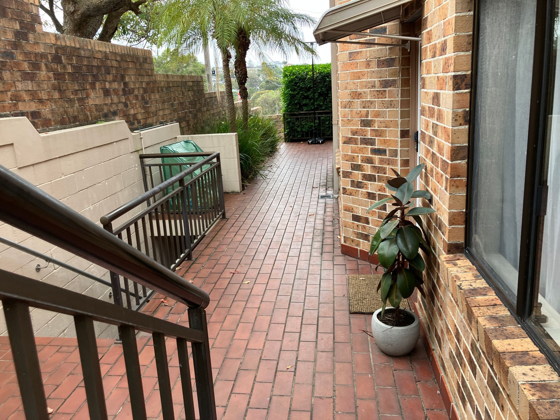 Brick patio walkway beside townhouses with railing, potted plant, and greenery at the end