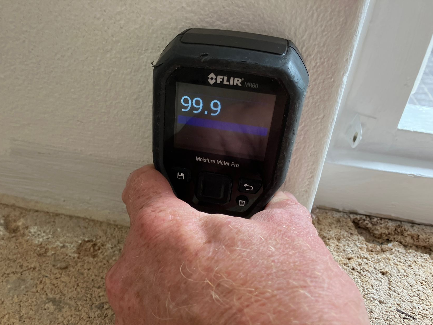 Hand holding a FLIR thermal camera aimed at a wall outlet, screen reading 99.9