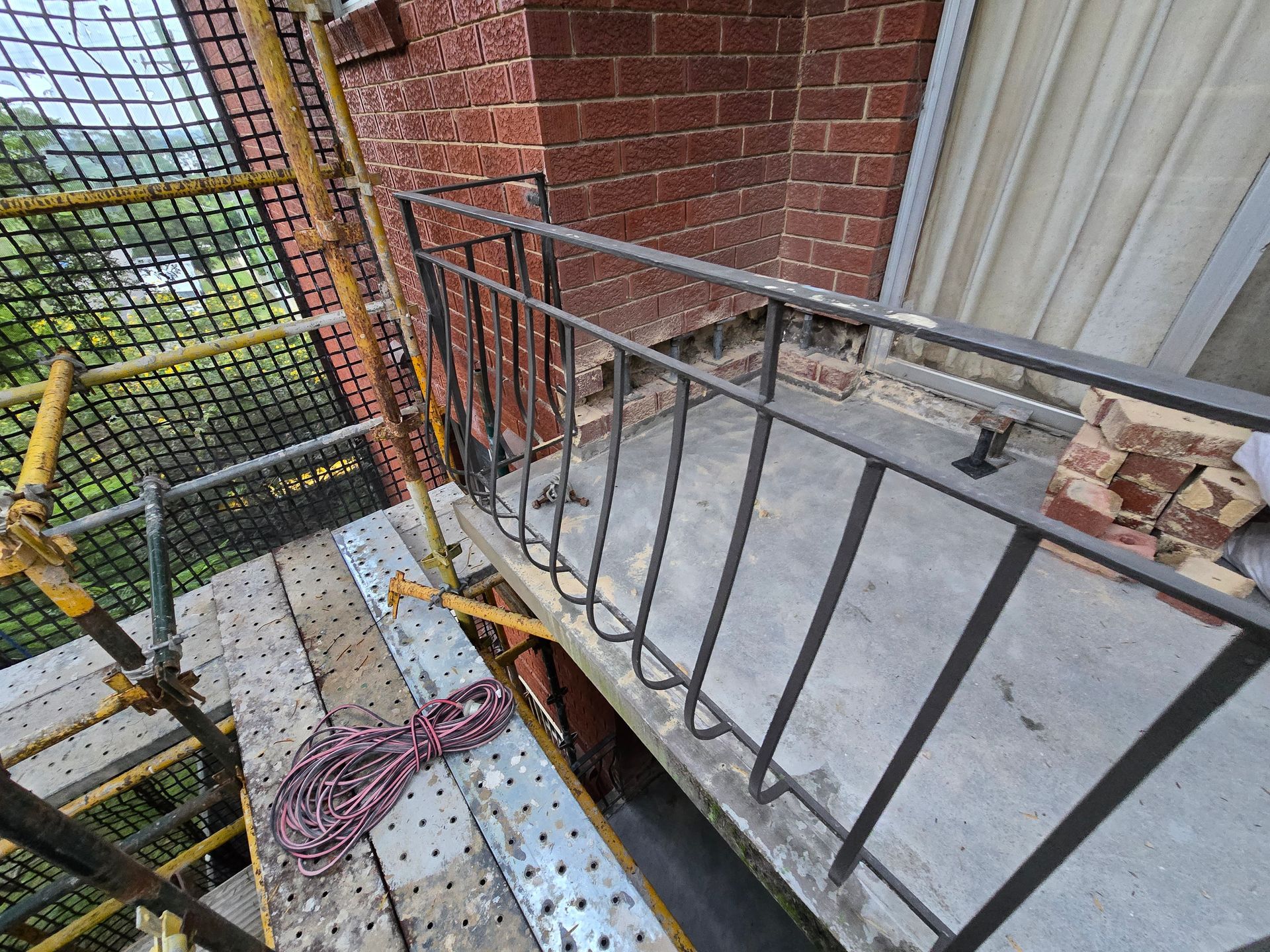 Scaffold walkway beside a brick wall, with metal railings, a window, and construction materials.