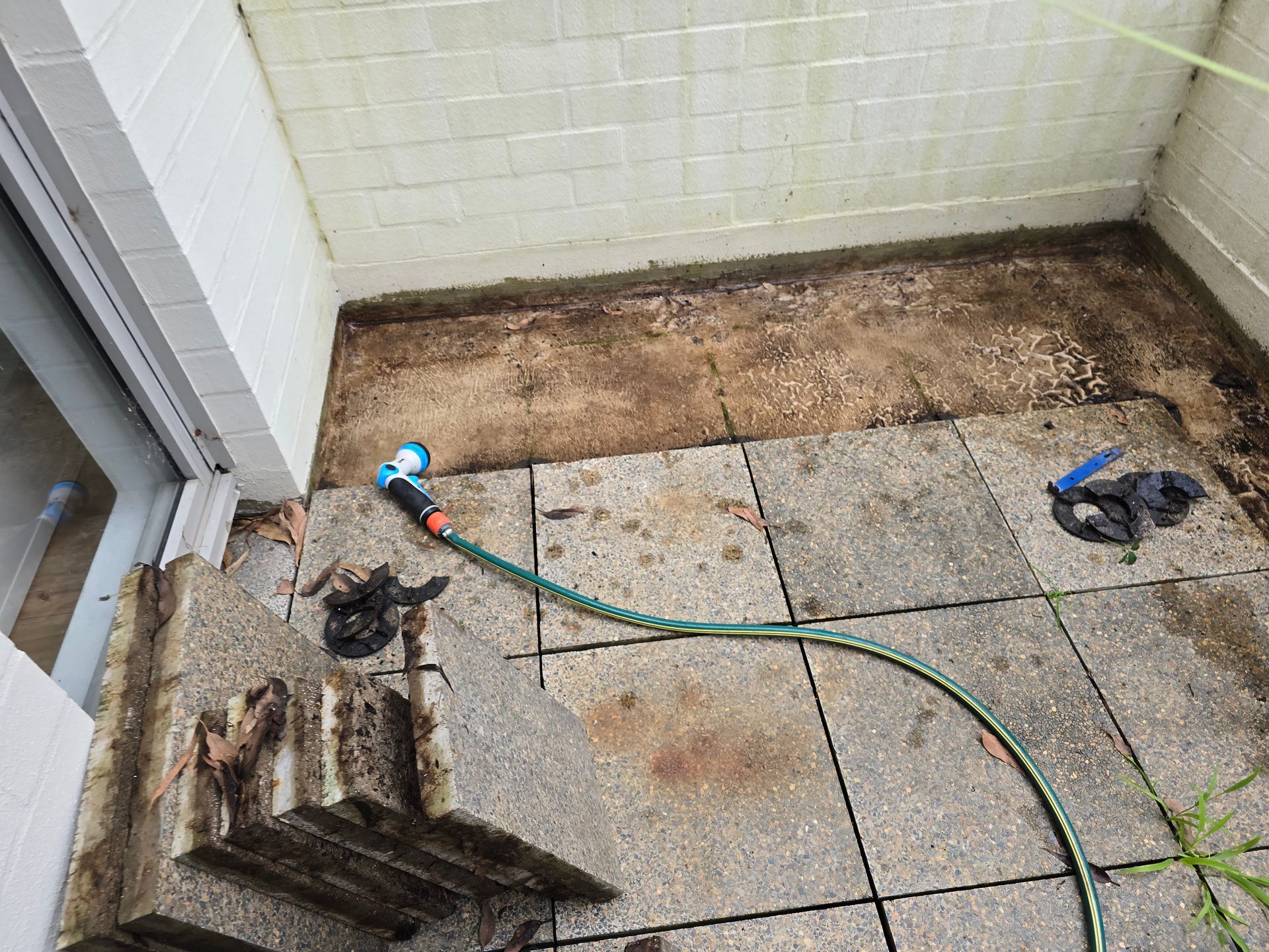 Small patio corner with pavers, dirt border, a hose, and a stack of wooden boards near a wall.