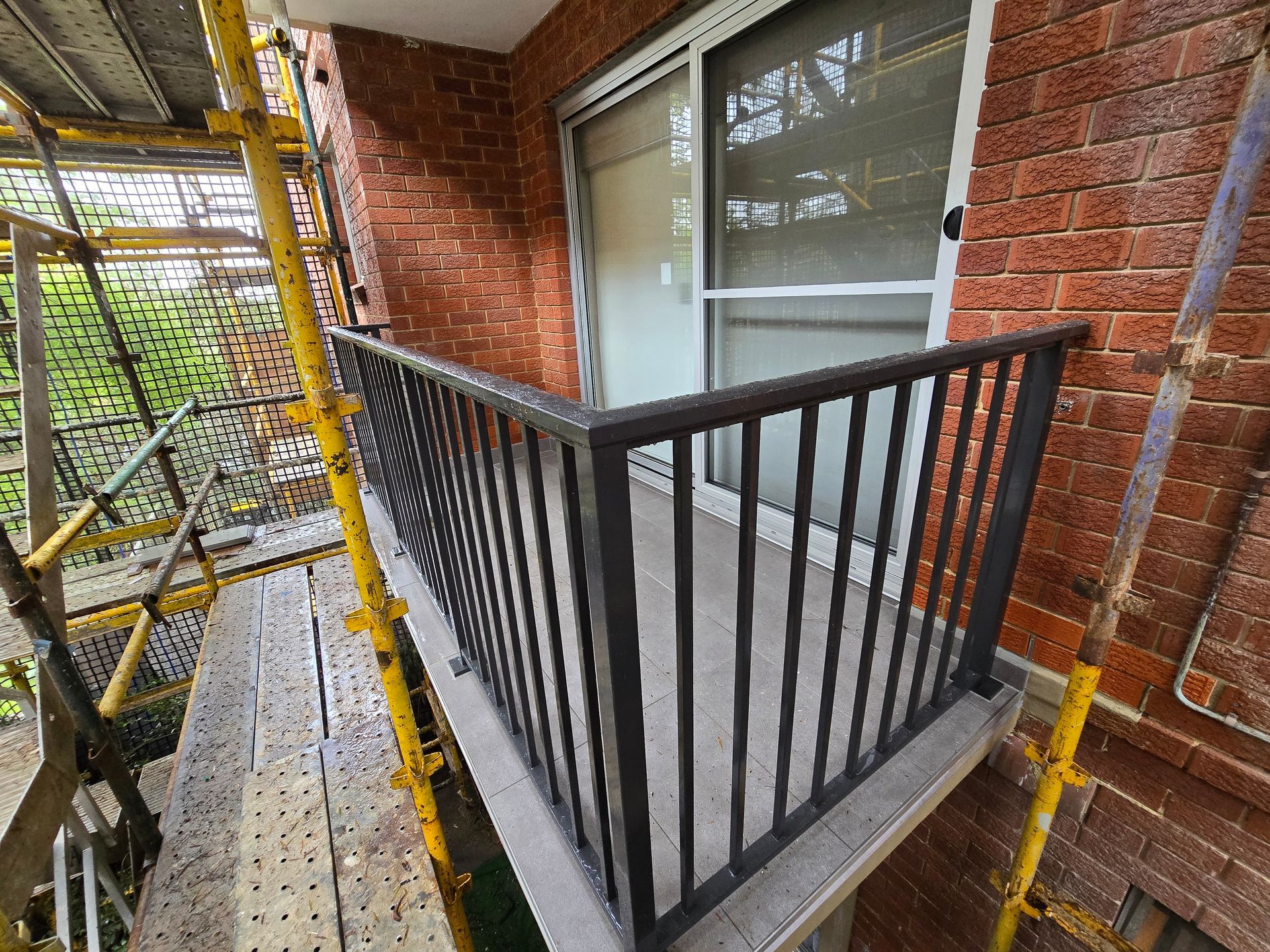 Narrow brick balcony with black railing, screened window, and scaffolding beside it