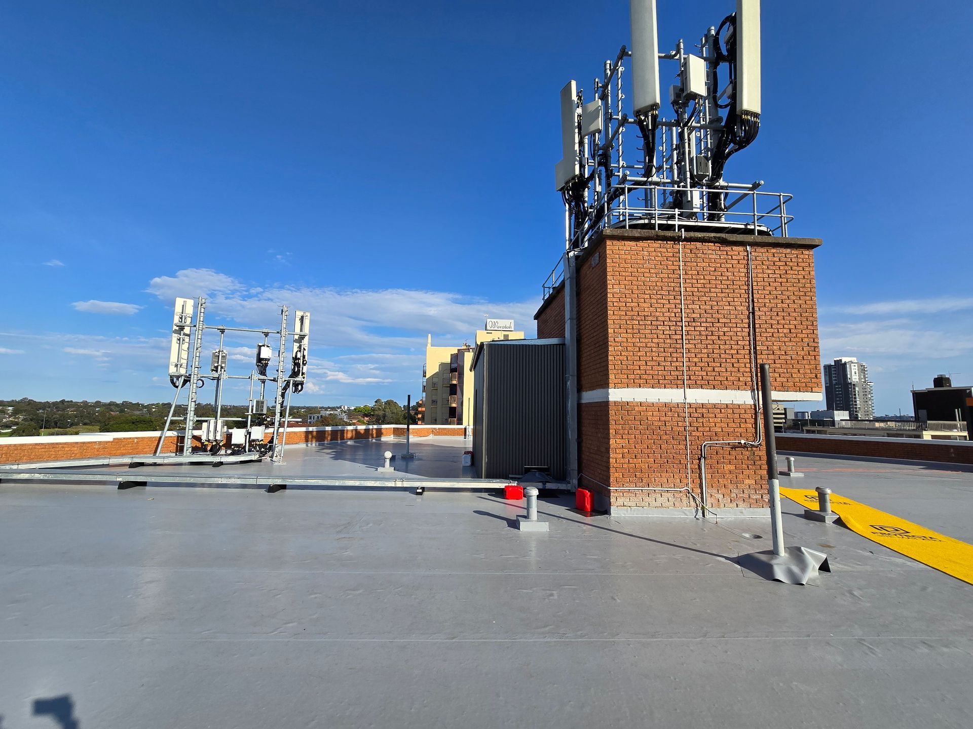 Rooftop with cellular antennas and equipment beside a brick utility structure under a blue sky