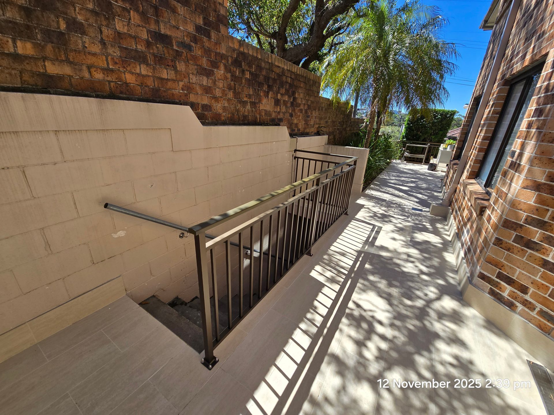 Outdoor brick walkway with metal railing beside a beige wall, bright sunlight and tree shadows.