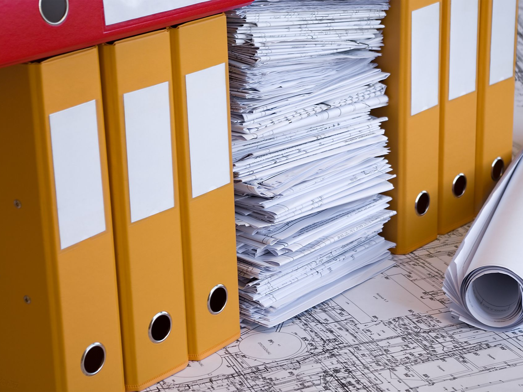 Yellow binders and a tall stack of papers on a desk, with a rolled blueprint nearby.