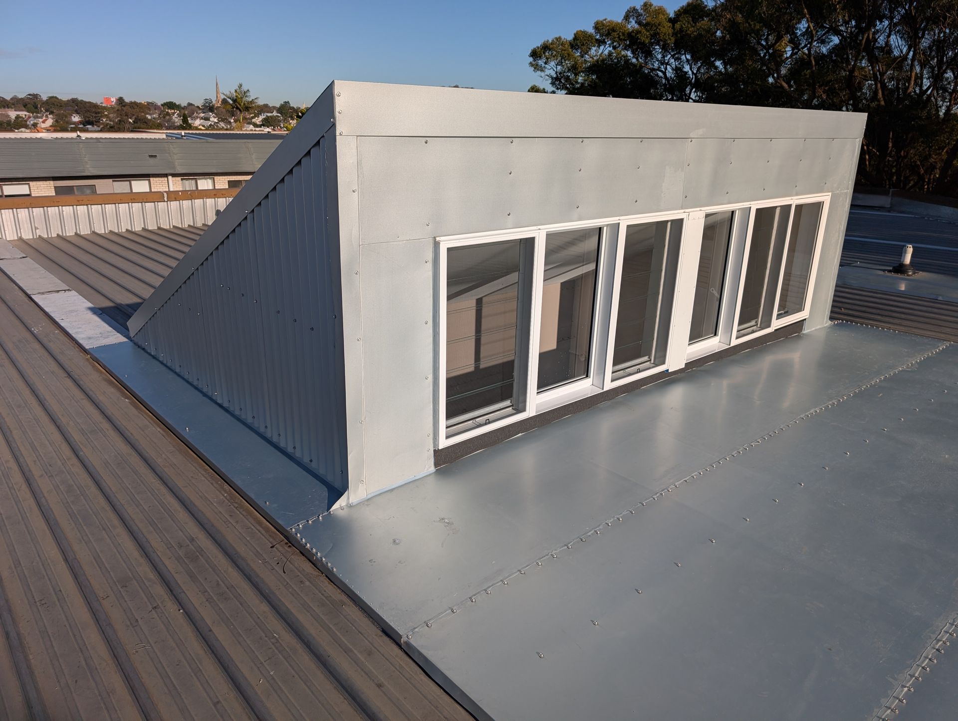 Rooftop dormer with white siding and windows on a metal roof under a clear blue sky