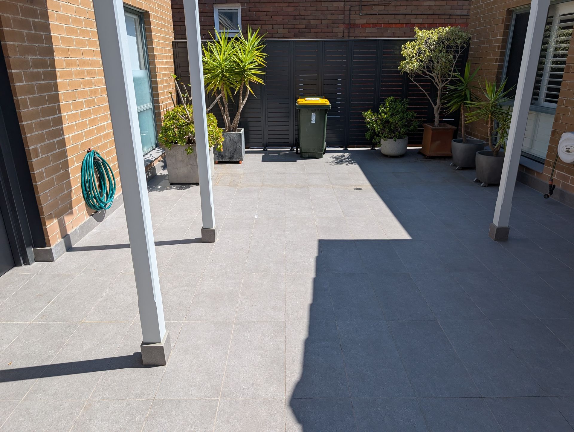 Outdoor patio with gray paving, white posts, potted plants, and trash bins against a brick wall