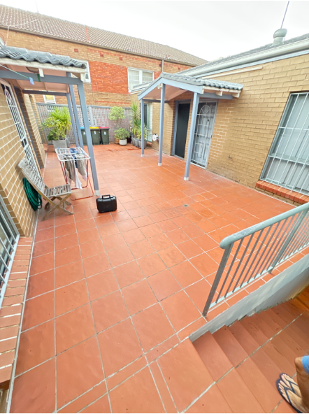 Terracotta-tiled courtyard between brick and tan buildings, with a small black bin and metal rails.