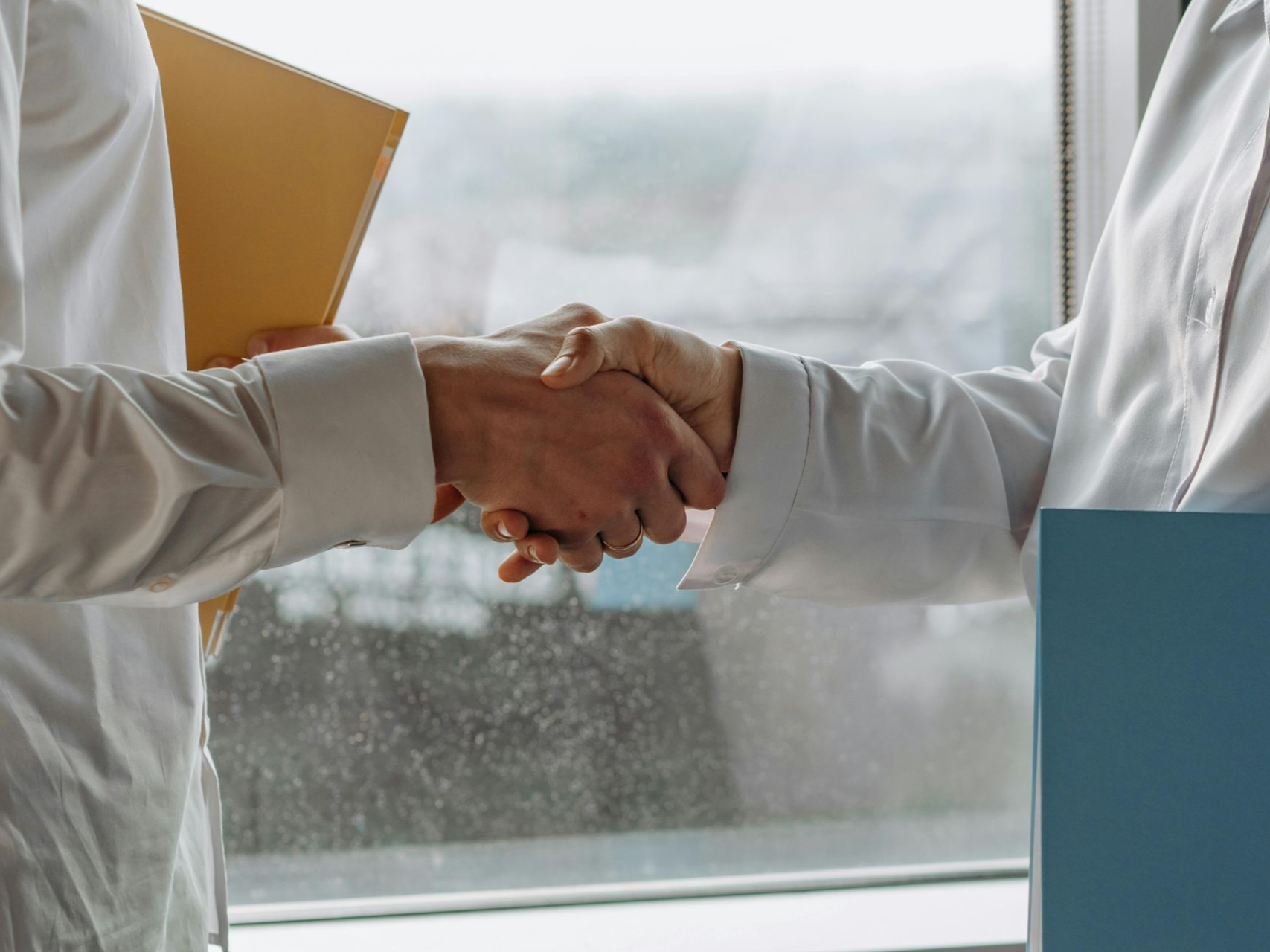Two people shake hands beside a window in an office, one holding a yellow folder.