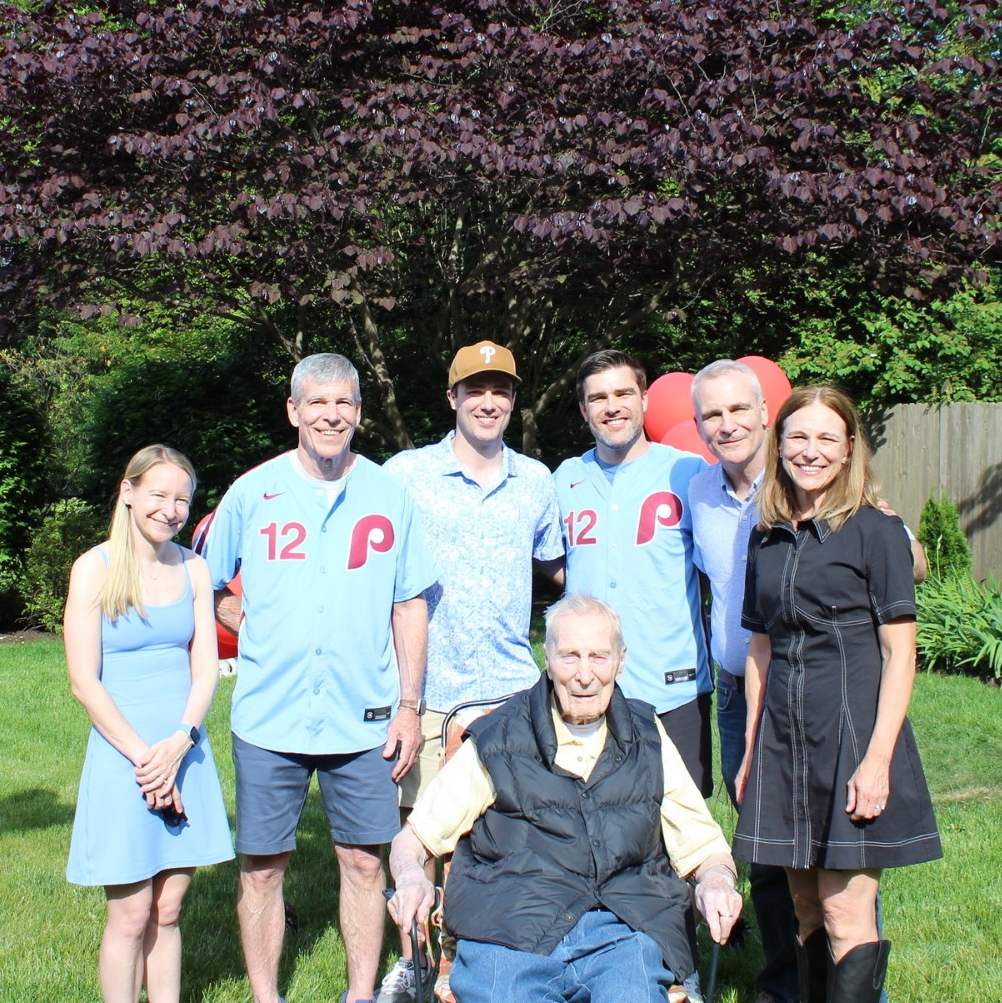 Three people standing in front of a house. A young woman and a man flank an elderly man, who is being supported. All are smiling.
