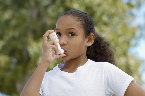 Young Black girl using an inhaler outdoors, holding it up to her mouth, with a worried expression.