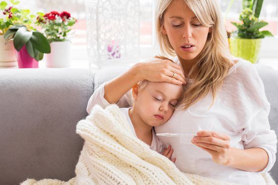 Woman checks child's temperature with thermometer. Child is wrapped in blanket, resting on mother's lap. Interior shot with plants in background.