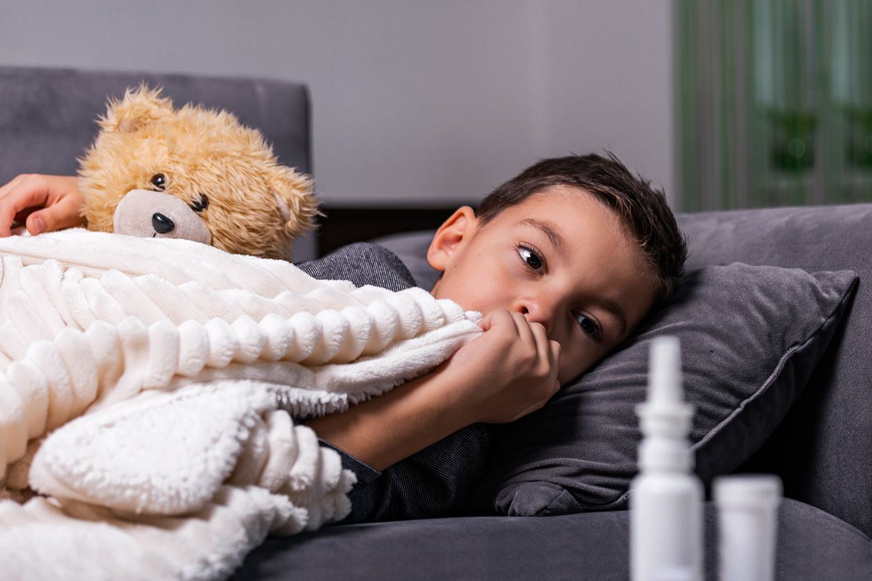 A young boy, looking unwell, lies on a couch under a blanket with a teddy bear. Nasal spray bottles sit nearby.