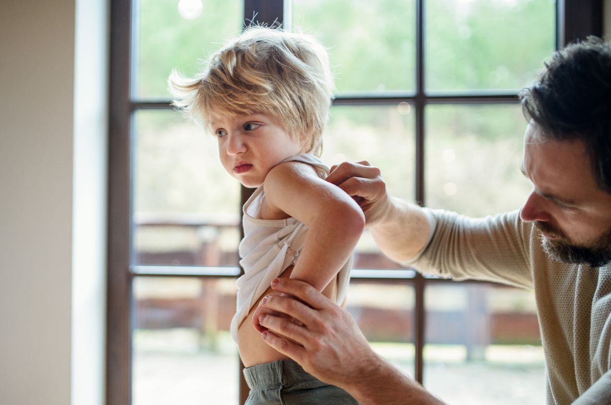A man examines a child's back, likely looking for a rash. They stand near a window. The child looks upset.