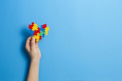 Hand holding a heart made of colorful puzzle pieces against a blue background, symbolizing autism awareness.