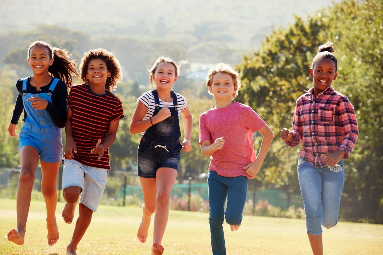 Five children running and smiling outdoors in a sunny park with a blurred background