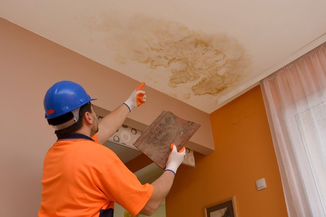 Person in blue helmet inspecting a water-damaged ceiling. Stained area in a room with orange walls.
