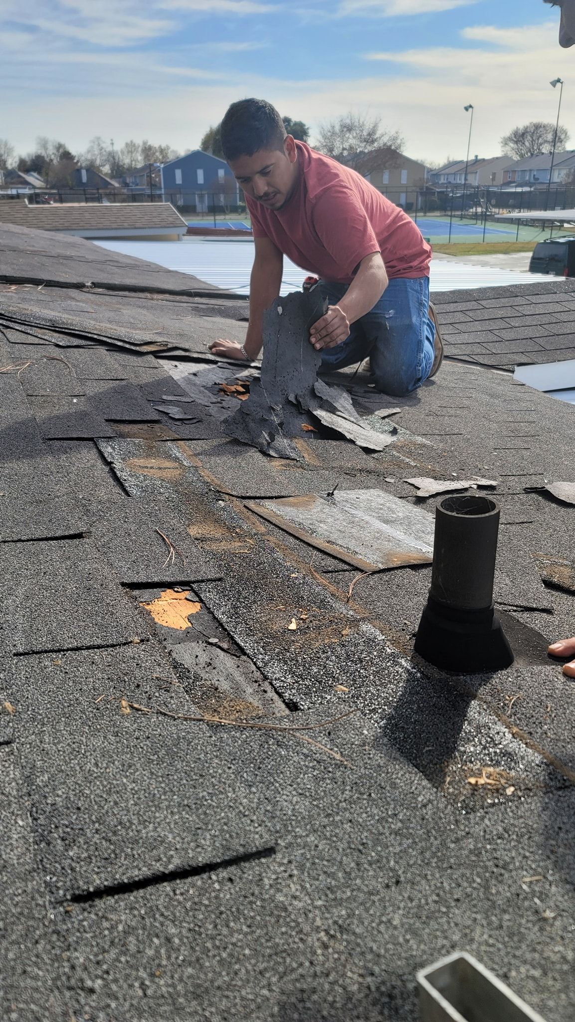 Worker kneeling on a flat roof, repairing damaged shingles near a vent in sunny weather.