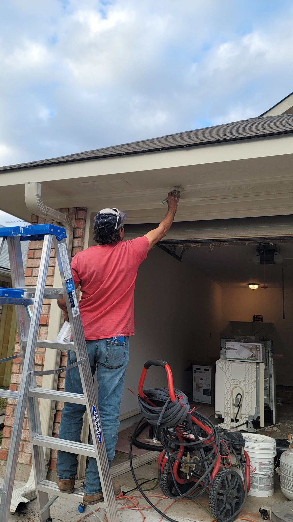 Person on a ladder painting the underside of a garage roof, with bikes and tools below.