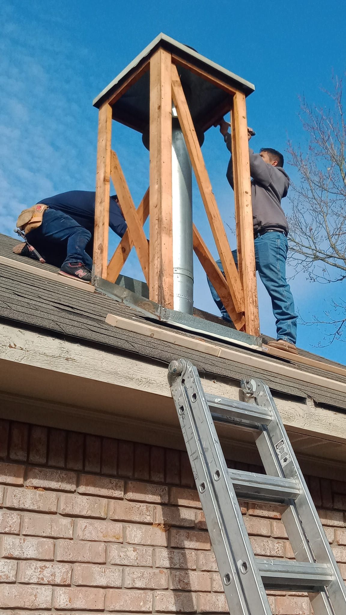 Workers installing a wooden chimney chase on a roof beside a ladder