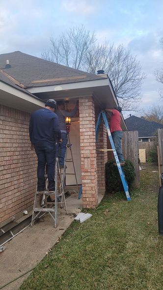 Two workers on ladders cleaning a brick house roof gutter beside a lawn