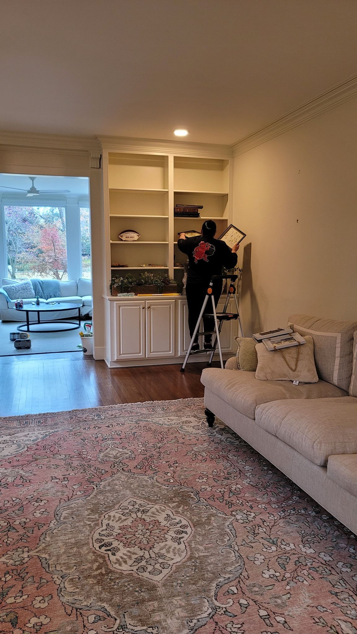 Cozy living room with beige sofa, patterned rug, built-in shelves, and a person standing near a lit alcove.