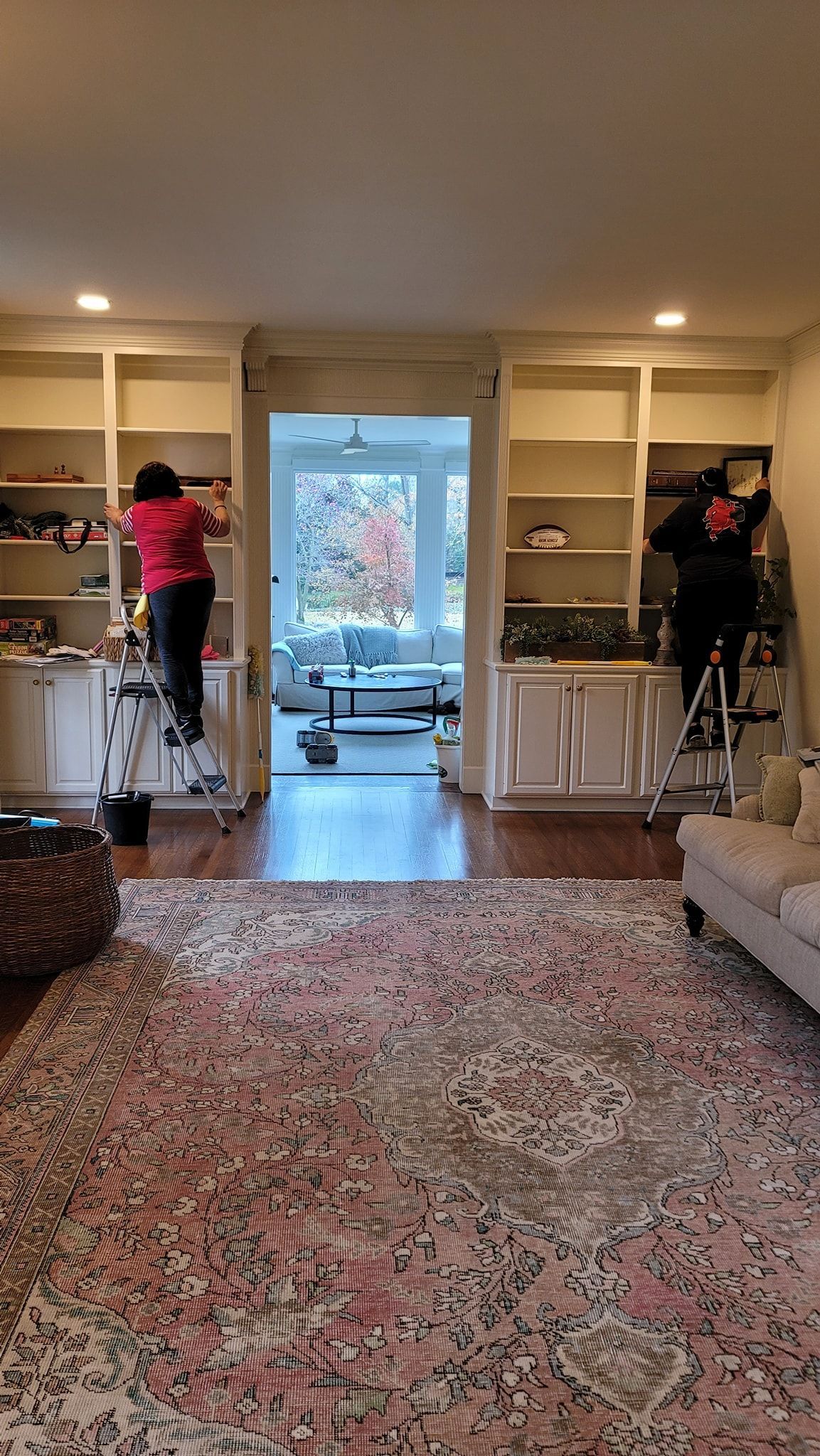 Living room with built-in shelves, a patterned rug, and two people on step ladders decorating the shelves.