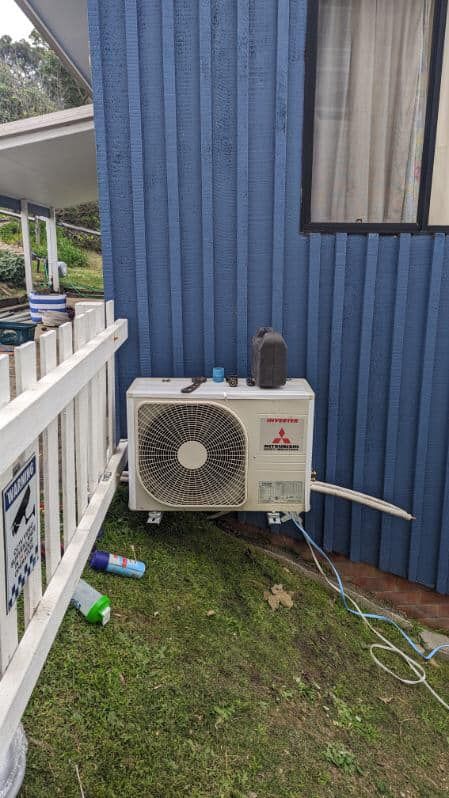 Outdoor air conditioner unit against a blue corrugated wall beside a white — Escarpment Air in Mount Saint Thomas, NSW