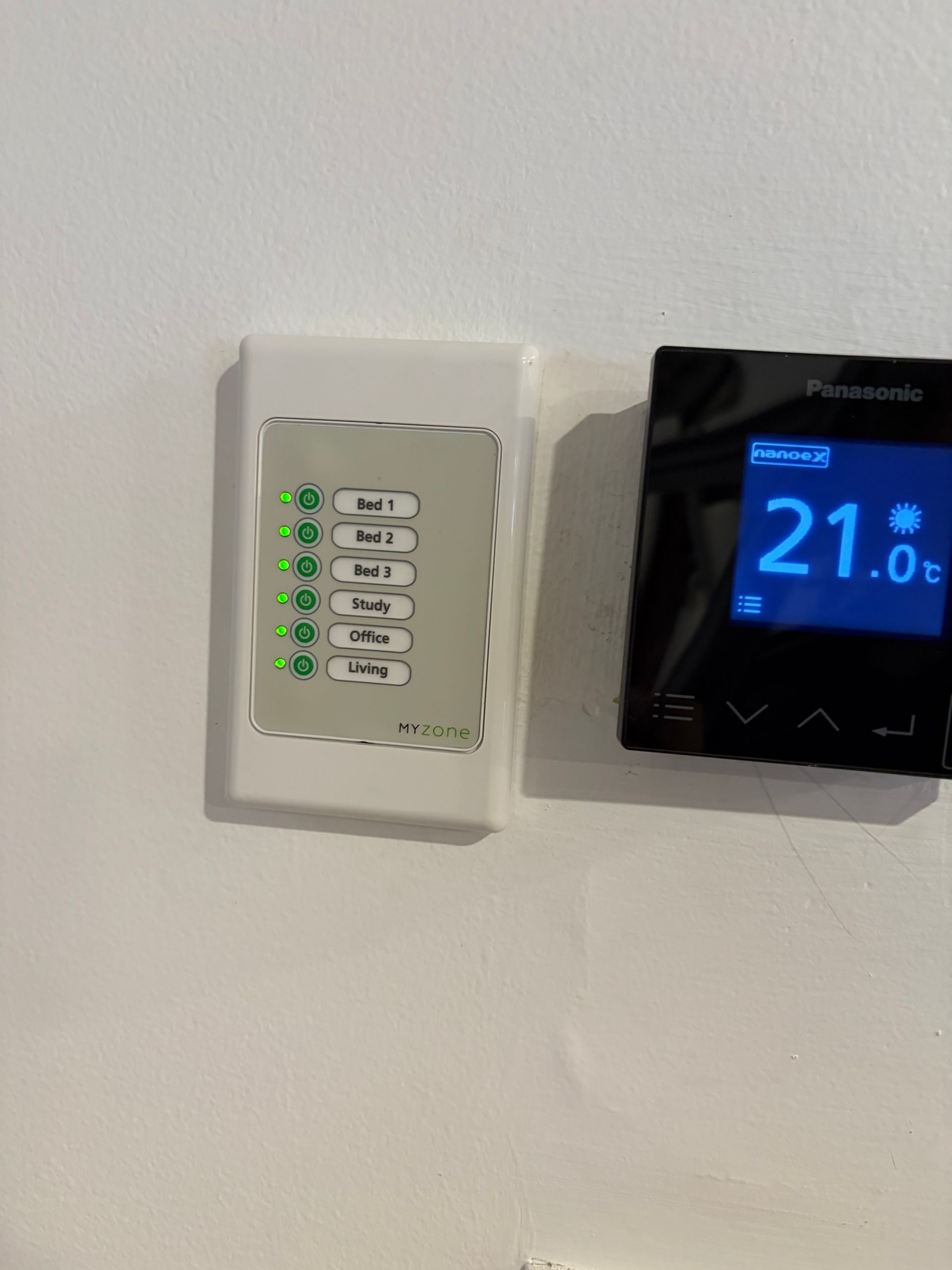 Wall-mounted thermostat with blue digital display beside a small white control panel with lit green buttons — Escarpment Air in Shellharbour, NSW